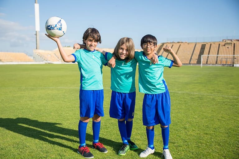 Three Children Standing on Green Grass Field With One Holding a Soccer Ball
